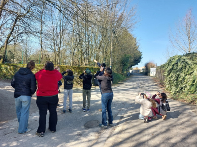 En pleine après midi ensoleillé, 6 adultes debout et 2 enfants accroupis accompagné d'un animateur LPO au milieu d'un rue d'une ville rurale observant avec une paire de jumelle chacun les oiseaux nichés dans les arbres alentours.